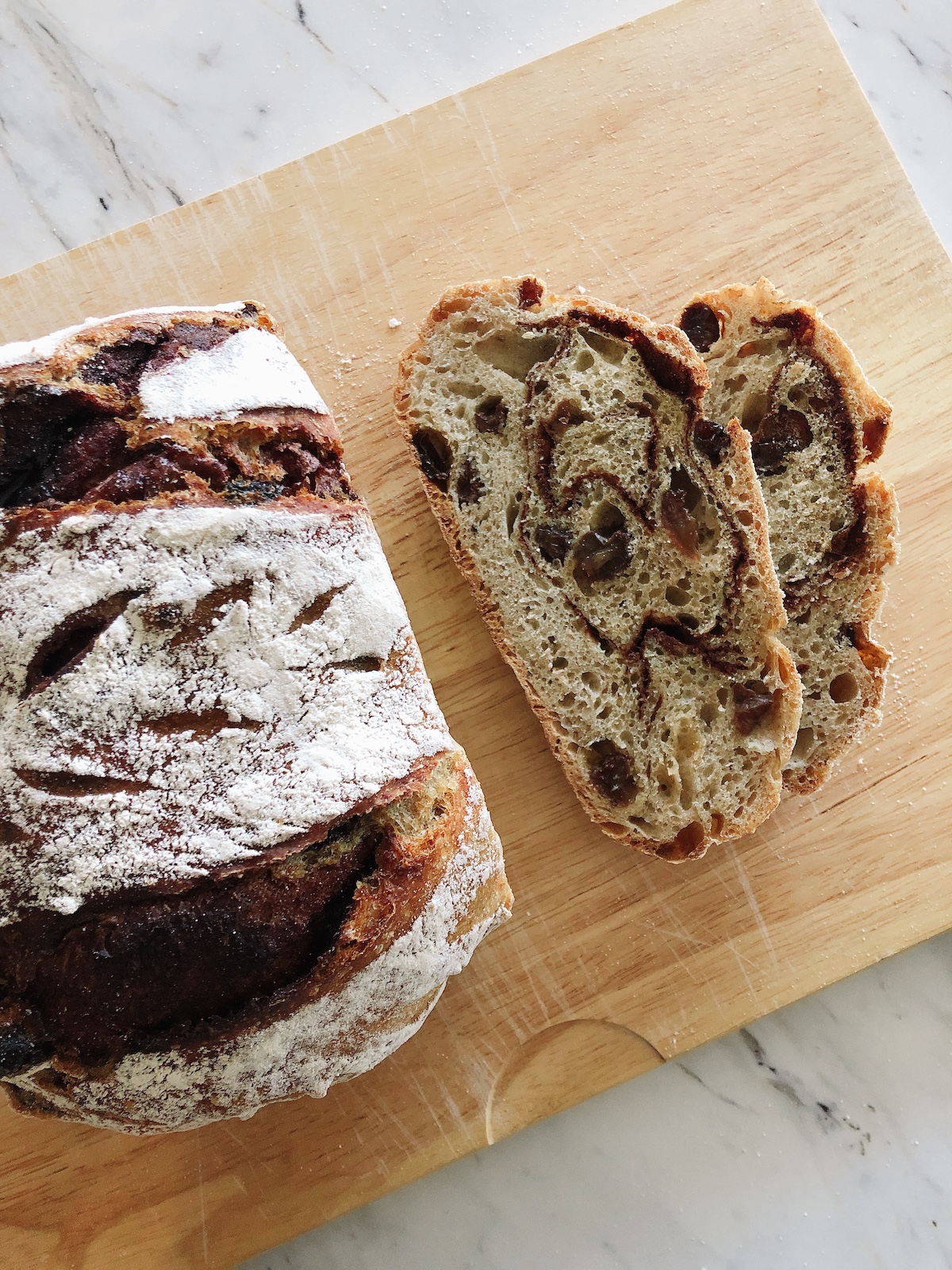 Two slices of cinnamon raisin no knead bread beside the entire loaf both on a wooden cutting board
