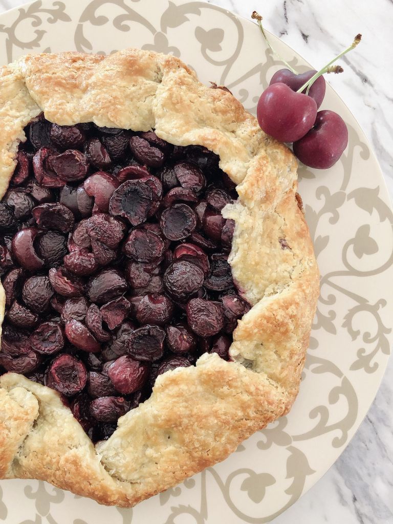 A close up of a rustic cherry galette on a decorative pie serving plate with three fresh cherries on the side