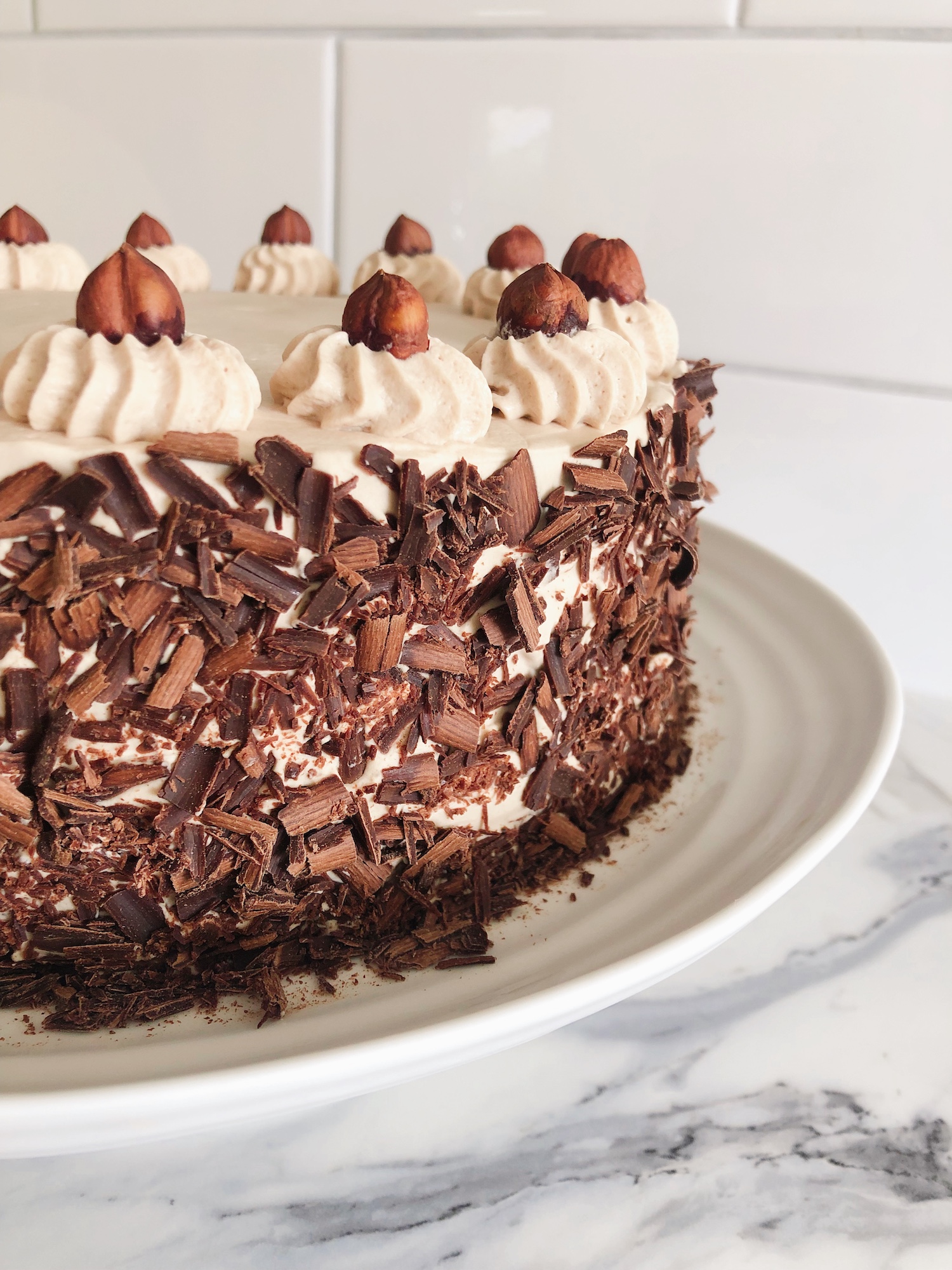 The side view of a Hazelnut Mocha Spelt Torte decorated with whole hazelnuts and chocolate shavings. The torte is on a low white cake stand with white tiles on the wall behind it.