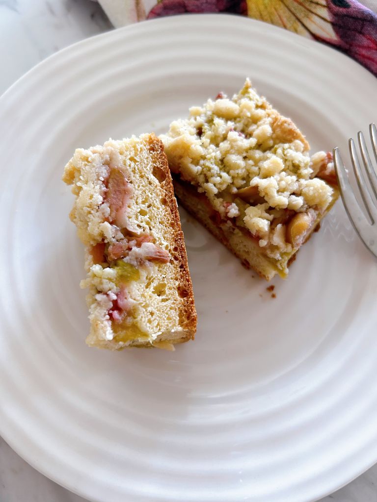 Two square slices of Once square slice of German Rhubarb Streusel Cake (Rhabarber-Streuselkuchen). Once slice is on its side showing the layers of streusel, rhubarb and cake. Both slices are sitting side by side on a small white plate.