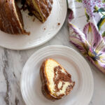 One slice of vanilla and chocolate marble cake on a white plate with the rest of the cake on a white cake plate beside it.