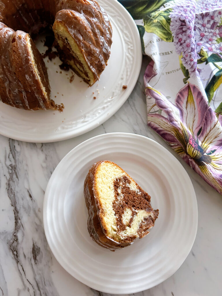 One slice of vanilla and chocolate marble cake on a white plate with the rest of the cake on a white cake plate beside it.