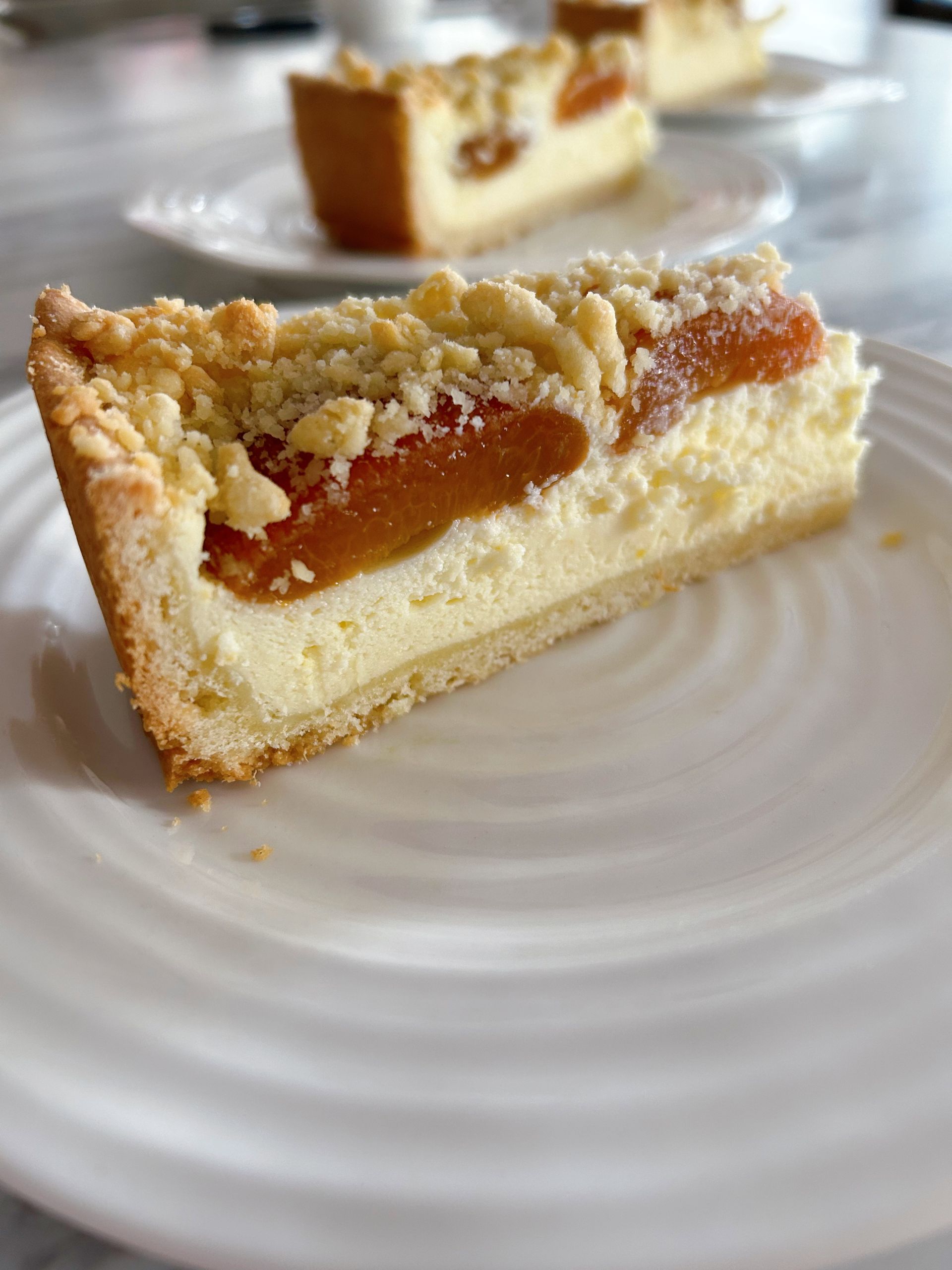 The side view of a single slice of German Apricot Cheesecake with Quark (Aprikosenkuchen) sitting on a white plate. Another slice is behind it also on a white plate.
