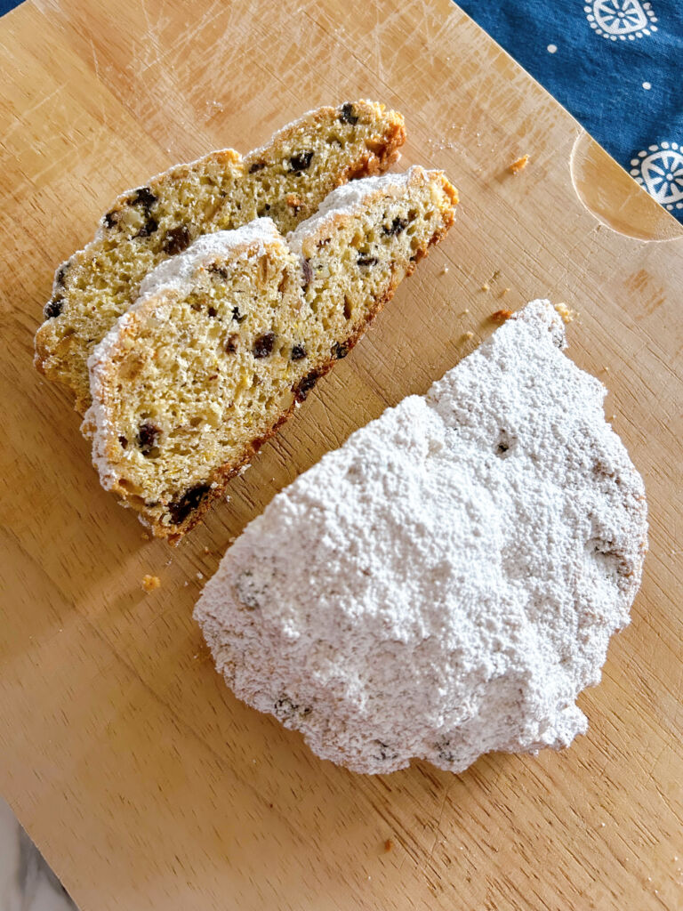 Two slices of German Christmas quark stollen overlapping each other on a wooden cutting board with the rest of the icing sugar dusted stollen close by.