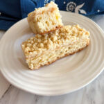Two slices of spelt strueselkuchen on a white plate with a blue and white tea towel in the background