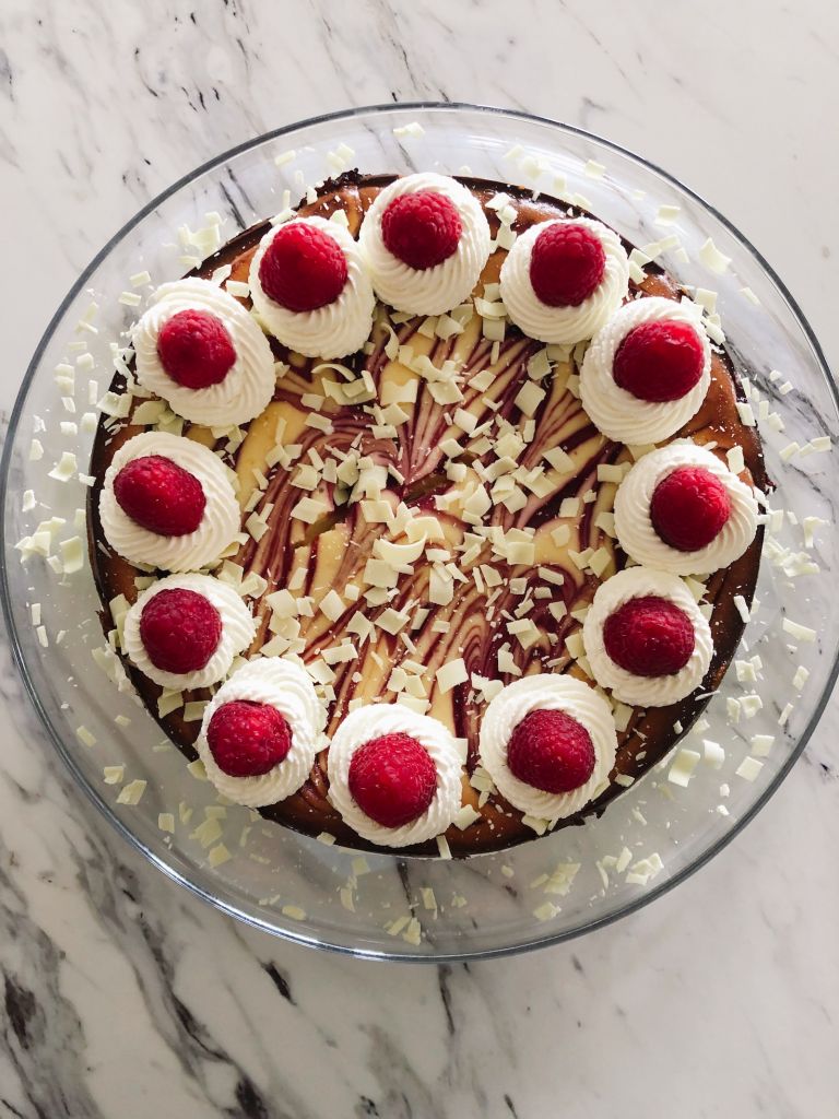 A round white chocolate cheesecake with raspberry swirl on a glass cake plate topped with twelve whipping cream swirls and a raspberry on top of each swirl