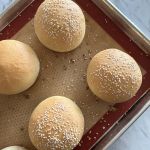 Four golden hued crust bread rolls topped with sesame seeds cooling on the lined baking sheet.