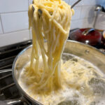 German Spätzle noodles being pulled out of a large pot of boiling water by a slotted pasta spoon.