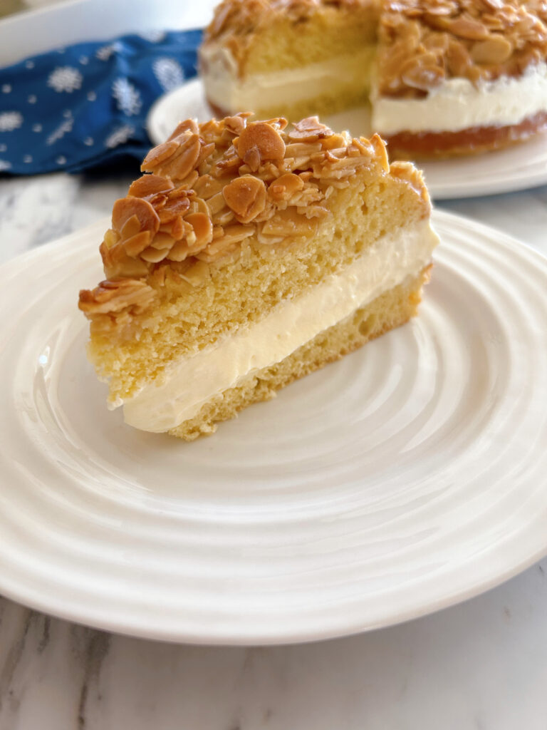 A side view of one slice of bee sting cake (aka Bienenstich) showing two layers of moist cake filled with a light and airy pastry and whipped cream and a toasted almond topping.