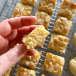 Small squares of dutch buttercake (Boterkoek) cooling on a wire rack while one is held above them between two fingers to show the dense texture.