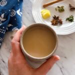 A large mug of masala chai with one hand wrapped around the mug. In the background is a white plate with spices on it and a dark blue and white tea towel.