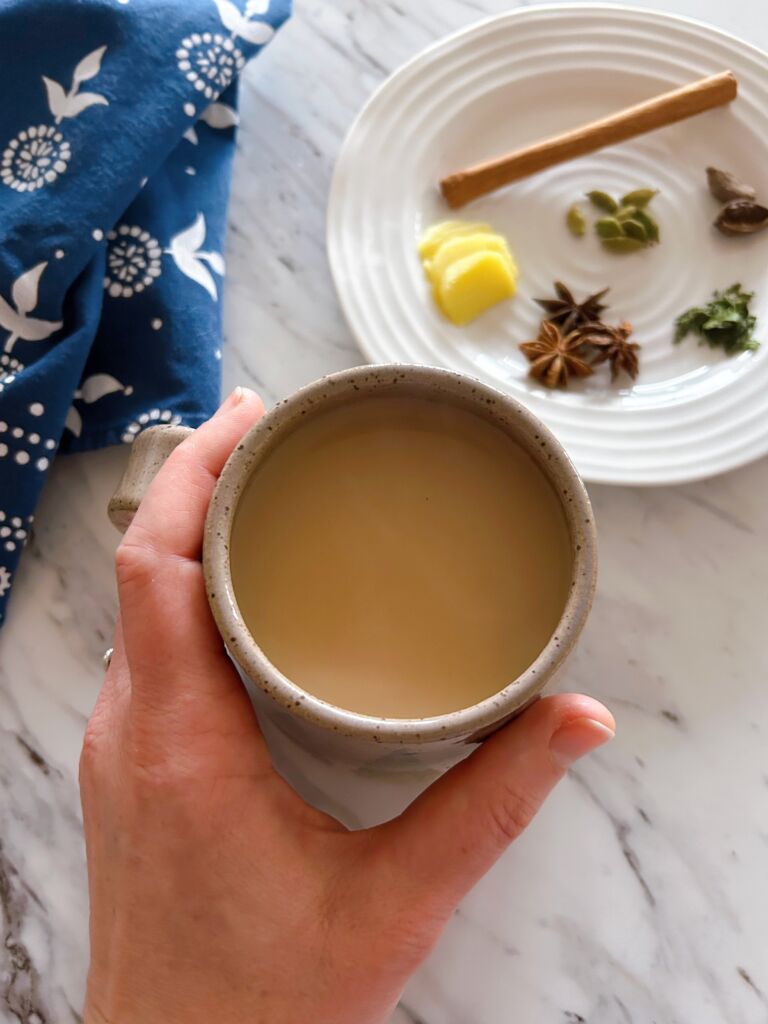 A large mug of masala chai with one hand wrapped around the mug. In the background is a white plate with spices on it and a dark blue and white tea towel.
